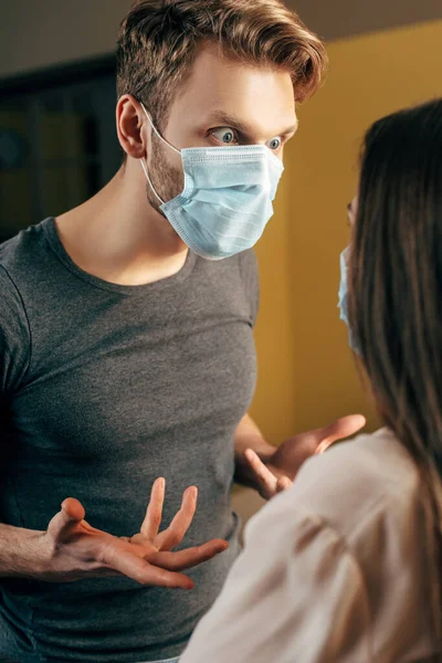Foco seletivo do homem emocional em gestos máscara médica enquanto briga com a namorada em casa — Fotografia de Stock
