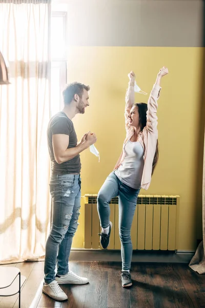 Mujer excitada sosteniendo máscara médica por encima de la cabeza y mirando al hombre guapo, fin del concepto de cuarentena - foto de stock