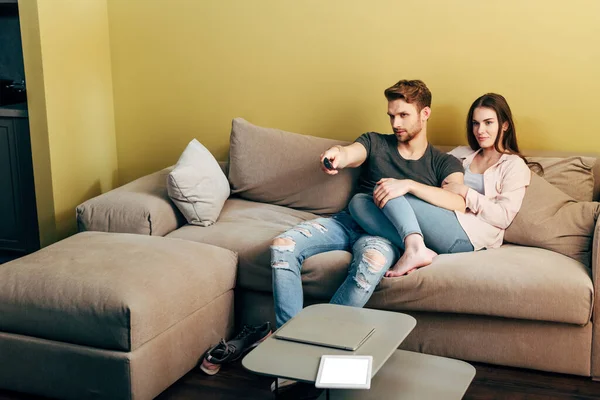 Bearded man watching movie near girlfriend, laptop and digital tablet with white screen on coffee table — Stock Photo