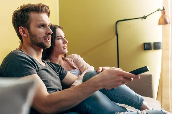 Selective focus of bearded man holding remote controller and watching movie with girlfriend at home — Stock Photo