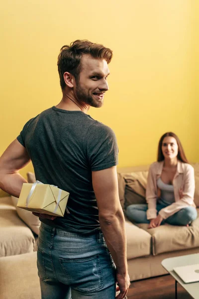 Selektiver Fokus des Mannes glücklich hält Geschenk hinter Rücken nahe Freundin im Wohnzimmer — Stockfoto