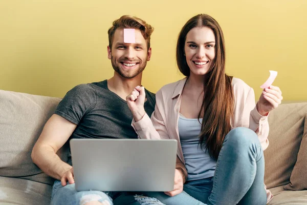 Alegre pareja con notas pegajosas en la frente jugando juego en línea cerca de la computadora portátil en casa - foto de stock