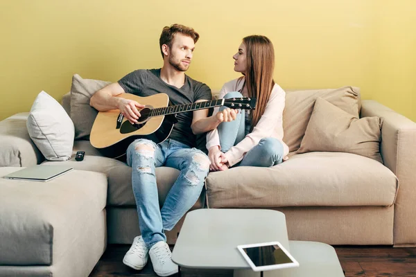 Guapo hombre tocando la guitarra acústica cerca de atractiva chica y tableta digital con pantalla en blanco - foto de stock