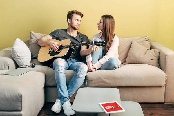 KYIV, UCRANIA - 22 DE ABRIL DE 2020: hombre guapo tocando la guitarra acústica cerca de chica atractiva y tableta digital con aplicación de youtube - foto de stock