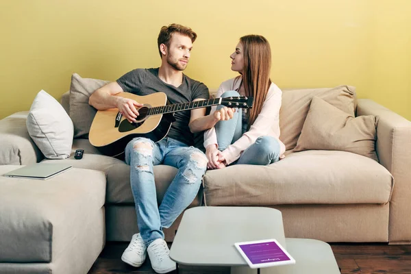 KYIV, UKRAINE - 22 AVRIL 2020 : bel homme jouant de la guitare acoustique près de fille attrayante et tablette numérique avec application instagram — Photo de stock