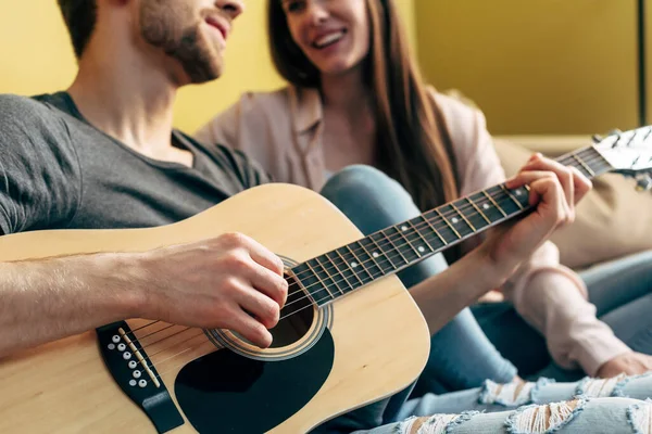 Обрізаний вид бородатого чоловіка, який грає на акустичній гітарі біля щасливої дівчини у вітальні — стокове фото