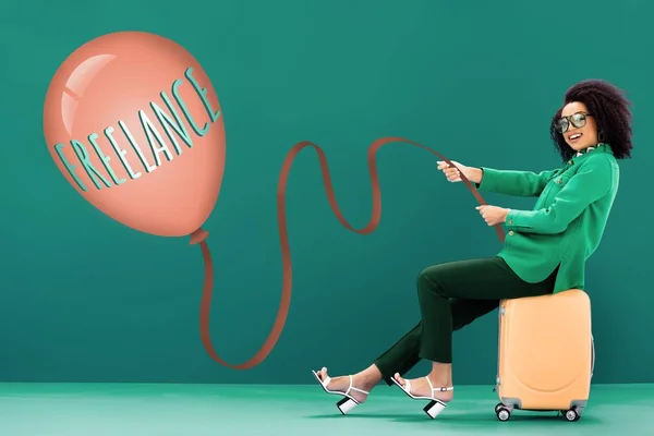 Smiling african american woman holding balloon with freelance lettering while sitting on travel bag on green background — Stock Photo