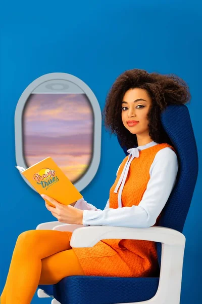 Femme afro-américaine souriante en robe rétro tenant livre avec des rêves deviennent réalité illustration tout en étant assis sur un siège isolé sur bleu avec hublot — Photo de stock