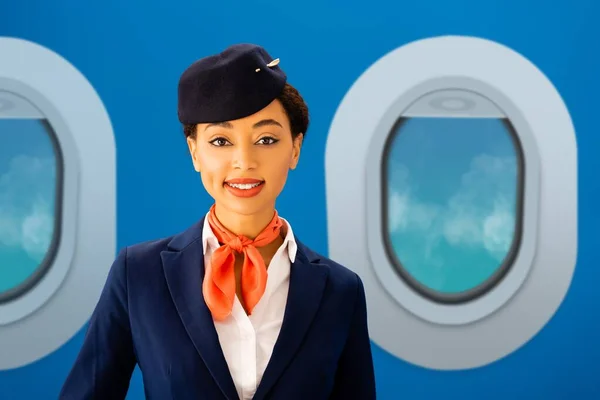 Smiling african american flight attendant looking at camera with portholes on blue background — Stock Photo