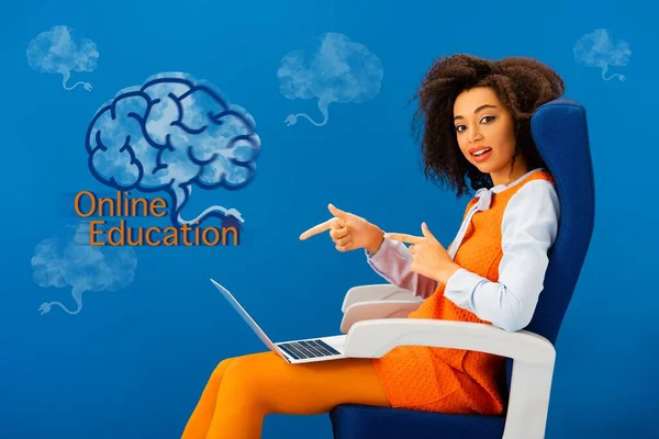 Donna afroamericana seduta sul sedile e che punta con le dita al computer portatile su sfondo blu con illustrazione educazione online — Foto stock