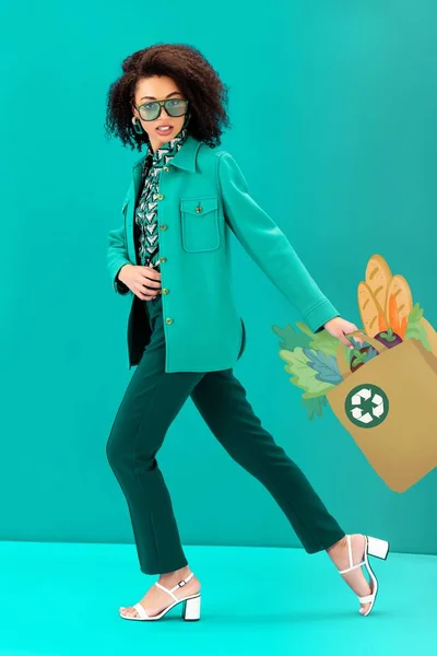 Stylish african american woman posing with paper bag with food and recycling sign on turquoise background — Stock Photo
