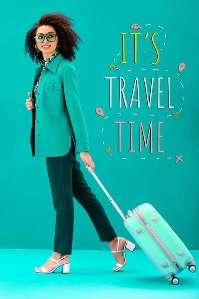 Smiling african american woman holding travel bag on turquoise background with it is travel time illustration — Stock Photo