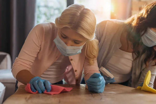 Selective focus of mother and daughter in medical masks cleaning table in living room — Stock Photo