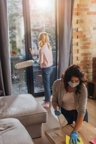 Selektiver Fokus der Frau in der medizinischen Maskenreinigungstabelle, während das Kind zu Hause Staub aufwirbelt — Stockfoto