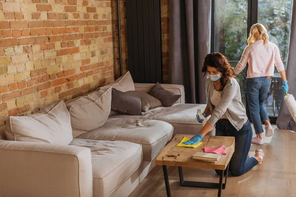 Mutter in medizinischer Maske putzt Couchtisch, während Tochter Staubbürste im Wohnzimmer hält — Stockfoto