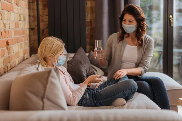 Selective focus of mother in medical mask holding glass of water near daughter with jar of pills on couch — Stock Photo