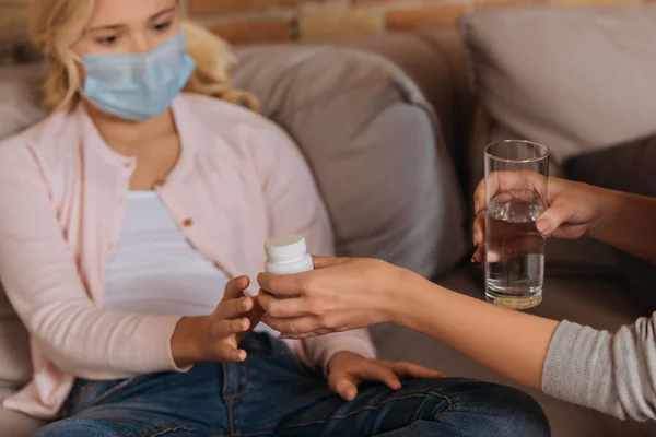 Focus selettivo della donna che dà vaso con pillole alla figlia in maschera medica sul divano — Foto stock