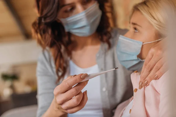 Selektiver Fokus der Mutter, die Thermometer in der Nähe des Kindes in medizinischer Maske hält — Stockfoto