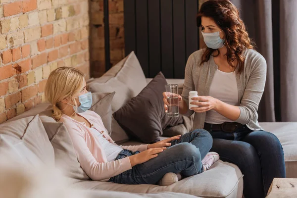 Focus selettivo della donna che tiene in mano un bicchiere d'acqua e un vaso con pillole vicino al bambino in maschera medica sul divano — Foto stock