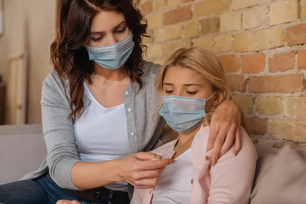 Selective focus of mother in medical mask holding thermometer near kid at home — Stock Photo