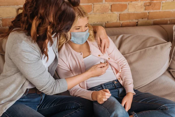 Woman holding thermometer near daughter in medical mask on couch — Stock Photo