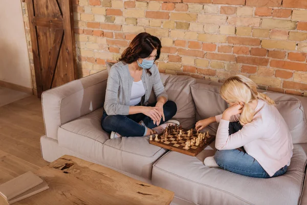 Мати в медичній масці грає в шахи з дочкою на дивані — стокове фото