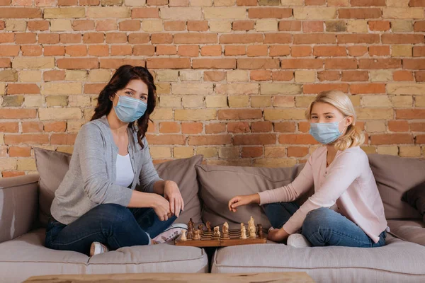 Vista lateral de la madre y la hija en máscaras médicas jugando ajedrez en el sofá - foto de stock