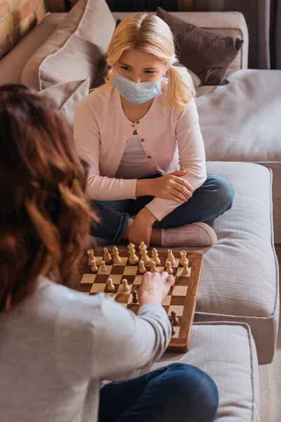 Focus selettivo del bambino in maschera medica che gioca a scacchi con la madre sul divano — Foto stock