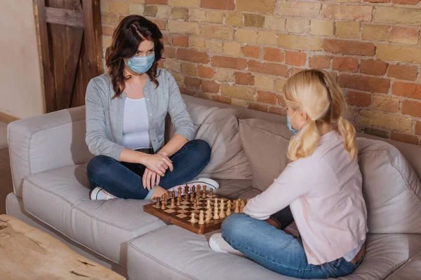 Жінка в медичній масці грає в шахи з дитиною, сидячи на дивані — стокове фото
