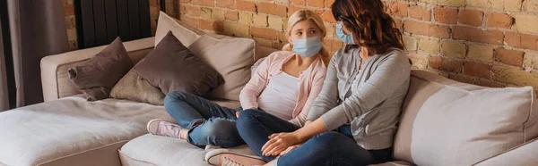 Panoramic orientation of mother and daughter in medical masks sitting on couch at home — Stock Photo