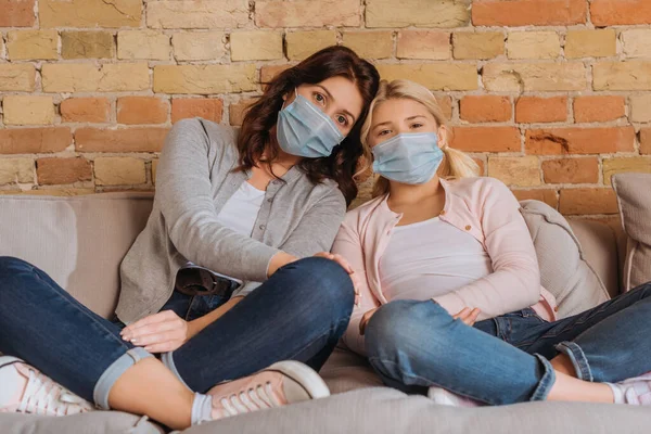 Madre e bambino in maschere mediche guardando la fotocamera sul divano a casa — Foto stock