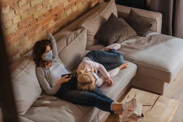 Visão de alto ângulo da mulher em máscara médica assistindo filme com a filha no sofá — Fotografia de Stock