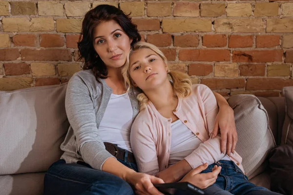 Woman holding remote controller and looking at camera while embracing daughter on couch — Stock Photo