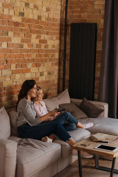 Vue latérale de la mère et de la fille souriantes regardant un film sur le canapé à la maison — Photo de stock