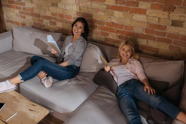 Vue grand angle de la mère et de la fille souriantes regardant la caméra tout en lisant des livres sur le canapé — Photo de stock