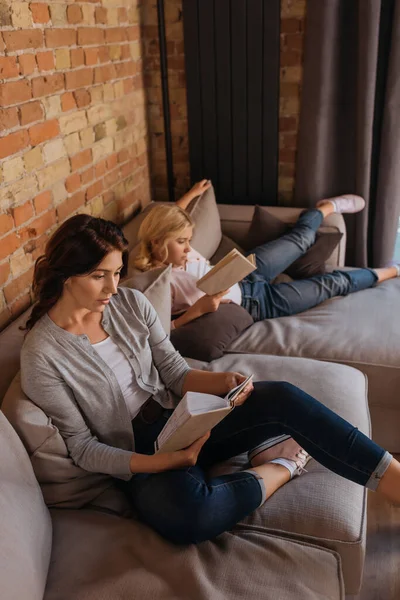 Madre e hija leyendo libros mientras están sentadas en el sofá en casa - foto de stock