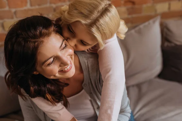 Ребенок целует улыбающуюся мать на диване дома — стоковое фото
