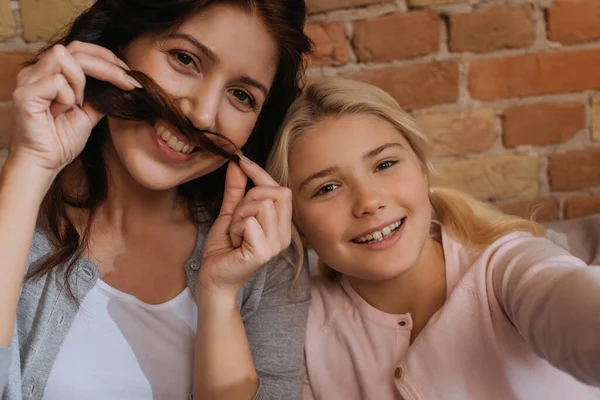 Mujer alegre y niño mirando a la cámara mientras sostiene el pelo cerca de la cara - foto de stock