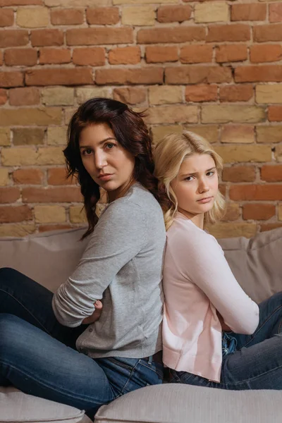 Side view of mother and daughter looking at camera while sitting back to back on couch — Stock Photo