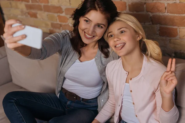 Выборочный фокус улыбающейся матери, делающей селфи со смартфоном рядом с дочерью, показывающей мир и торчащей языком — стоковое фото