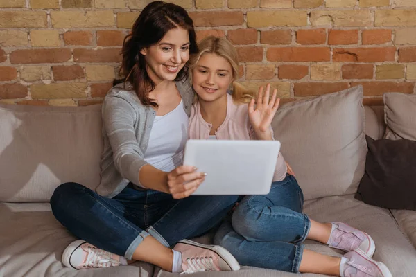 Selective focus of smiling kid and mother having video call on digital tablet on couch — Stock Photo
