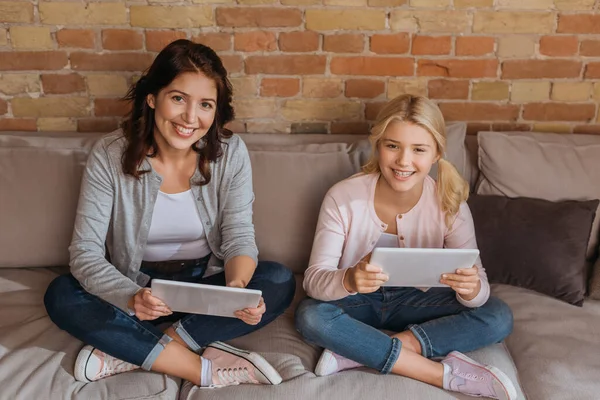 Lächelnde Mutter und Kind blicken in die Kamera, während sie digitale Tablets auf der Couch halten — Stockfoto