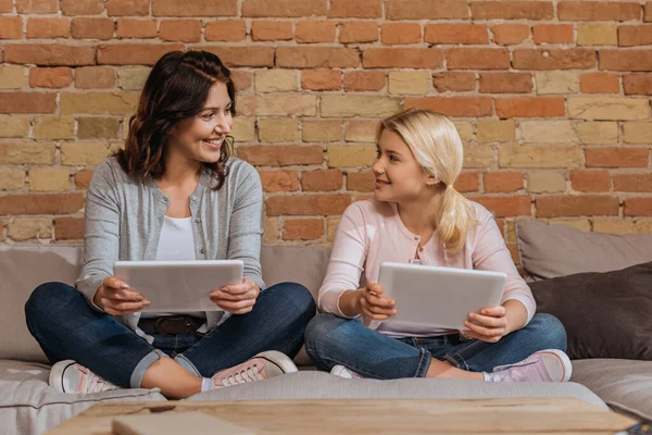 Enfoque selectivo de sonreír madre e hija sosteniendo tabletas digitales en el sofá - foto de stock