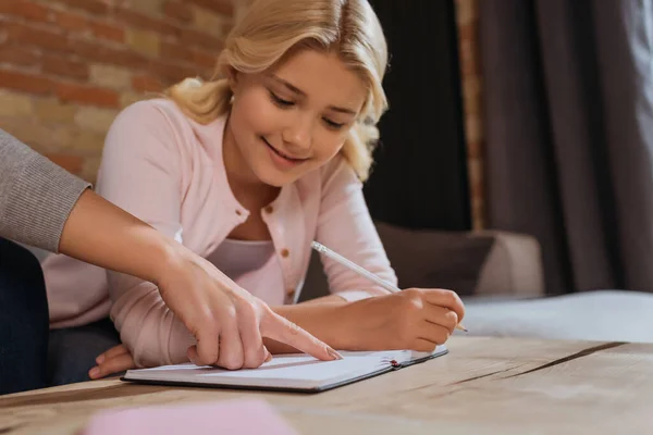 Enfoque selectivo del niño sonriente sosteniendo el lápiz cerca de la madre señalando el cuaderno en casa - foto de stock