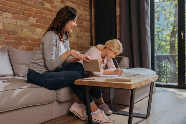 Madre sonriente sosteniendo libro cerca de niño escribiendo en el cuaderno en la mesa de café en la sala de estar - foto de stock