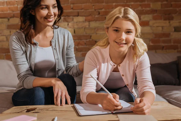 Enfoque selectivo del niño sonriente mirando la cámara mientras escribe en el cuaderno cerca de la madre en el sofá - foto de stock