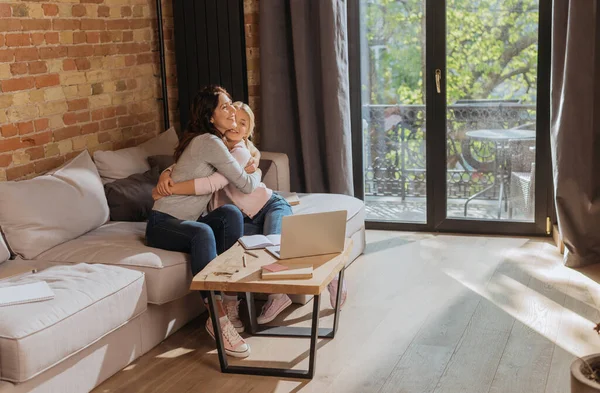 Mère souriante étreignant l'enfant près de l'ordinateur portable, ordinateur portable et livre sur la table basse à la maison — Photo de stock