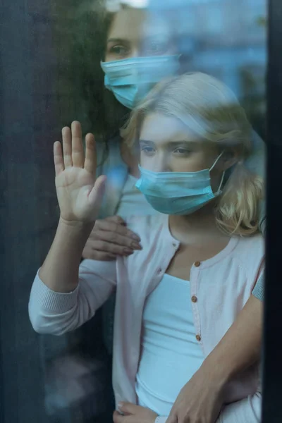 Selective focus of mother in medical mask embracing child near window at home — Stock Photo
