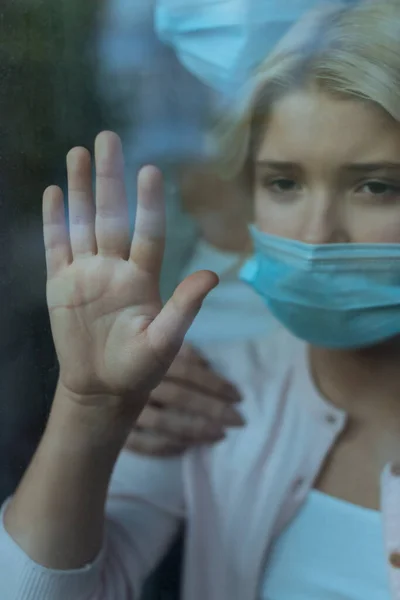 Selective focus of kid in medical mask touching galas of window near mother — Stock Photo