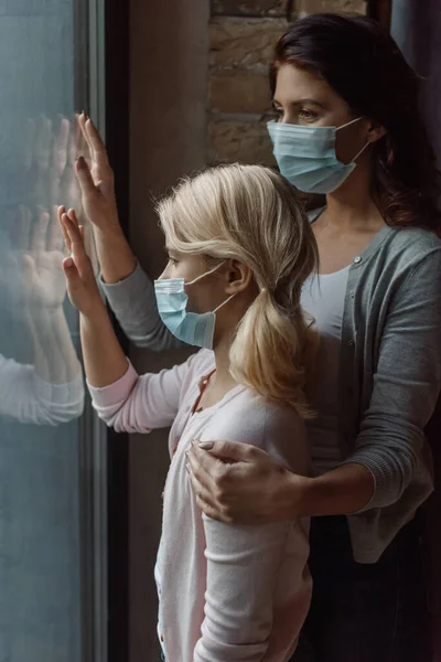 Мать в медицинской маске обнимает дочь у окна дома — стоковое фото